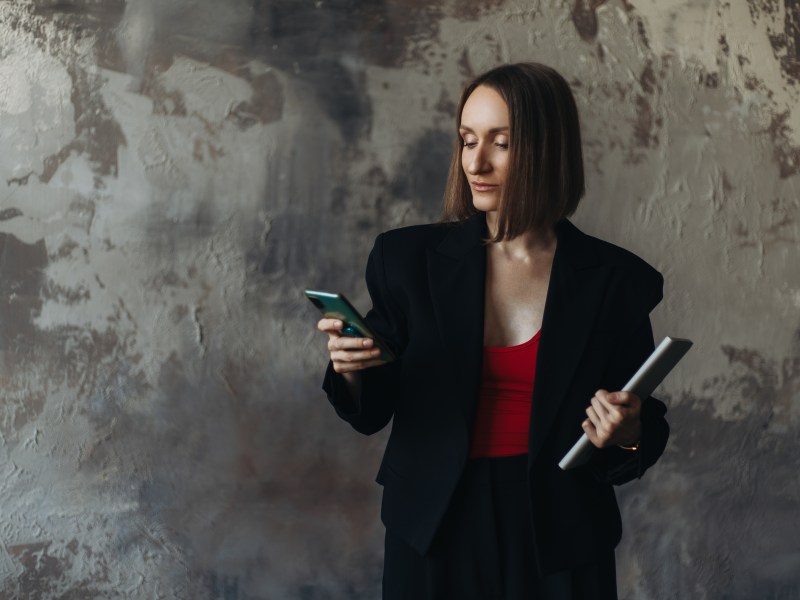 Dreiviertelaufnahme einer GeschÃ¤ftsfrau in einer schwarzen Jacke, die ihr Smartphone benutzt und ein Tablet in der Hand hÃ¤lt.