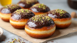 6 Dubai-Schokolade-Donuts mit Pistazien bestreut und Schokolade Ã¼berzogen auf Backpapier.