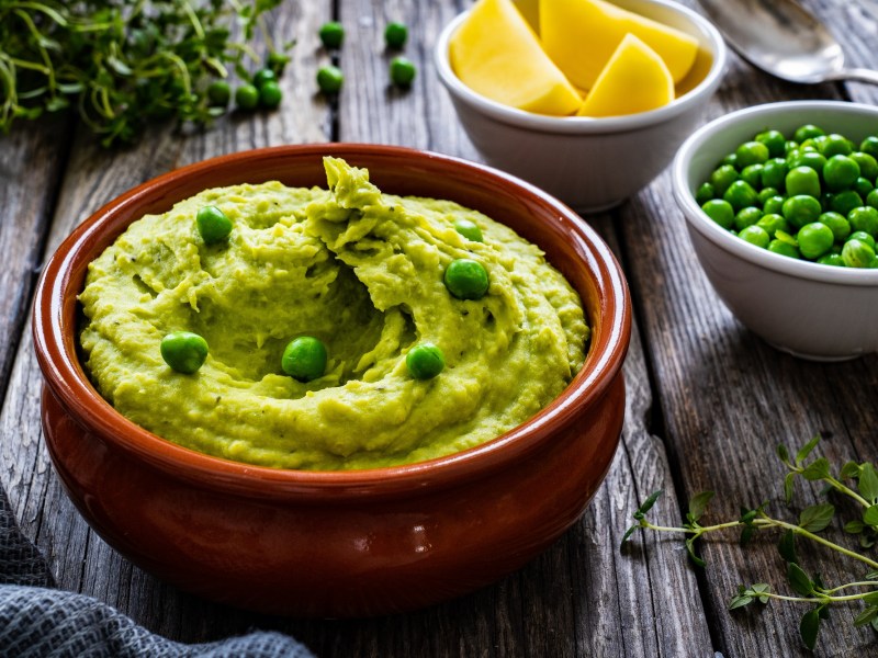 Eine braune Schale Erbsen-Guacamole, garniert mit ganzen Erbsen, daneben eine Schale Erbsen und eine Schale Zitronenspalten sowie KrÃ¤uter.