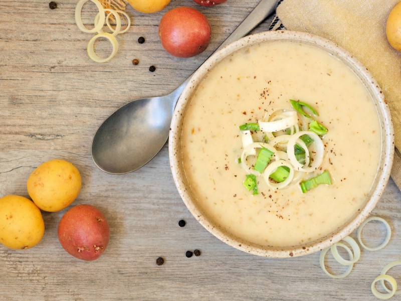 Eine SchÃ¼ssel Kartoffel-Lauch-Suppe mit Lauch und Pfeffer garniert, daneben ein LÃ¶ffel und Kartoffeln, Draufsicht.