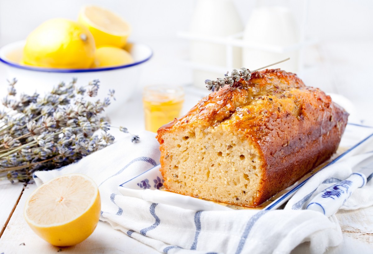 ein angeschnittener franzÃ¶sischer Lavendelkuchen auf einem karierten Geschirrtuch, daneben Lavendel und Zitronen.