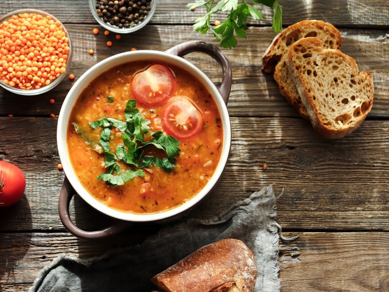 Eine SchÃ¼ssel mit Henkeln, darin Linsen-Tomaten-Suppe, garniert mit halbierten Tomaten und Koriander, daneben frische Tomaten, Brotscheiben, eine Schale Linsen, Draufsicht.