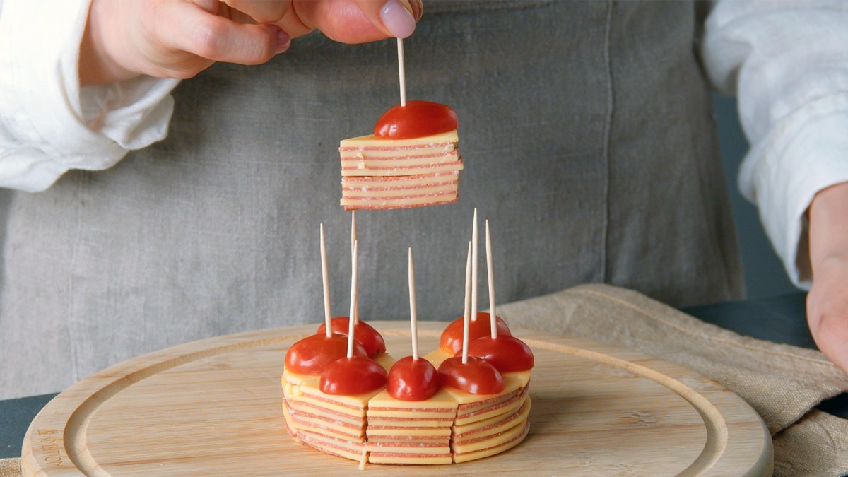 Salami-KÃ¤se-Snack mit Tomate auf einem Holzbrett. Die Hand einer KÃ¶chin hÃ¤lt ein StÃ¼ck in die Kamera