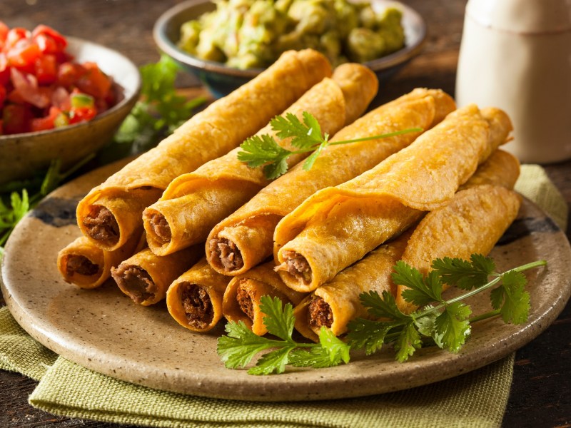 Taquitos mit HackfleischfÃ¼llung auf einem beigen Teller, garniert mit Koriander und serviert mit Tomaten-Salsa, Guacamole und Sour Cream.