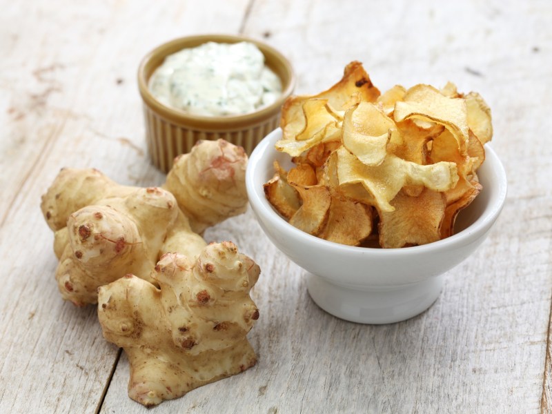 Eine Schale Topinambur-Chips. Daneben liegen ganze Knollen und ein Dip.