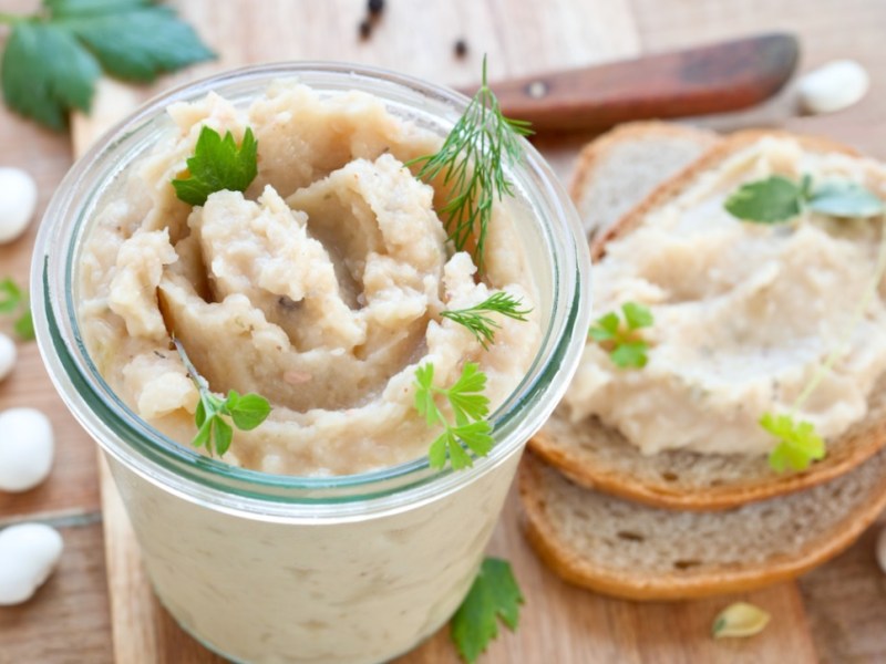 WeiÃŸe-Bohnen-Aufstrich im Glas. Daneben eine Scheibe Brot mit Aufstrich.