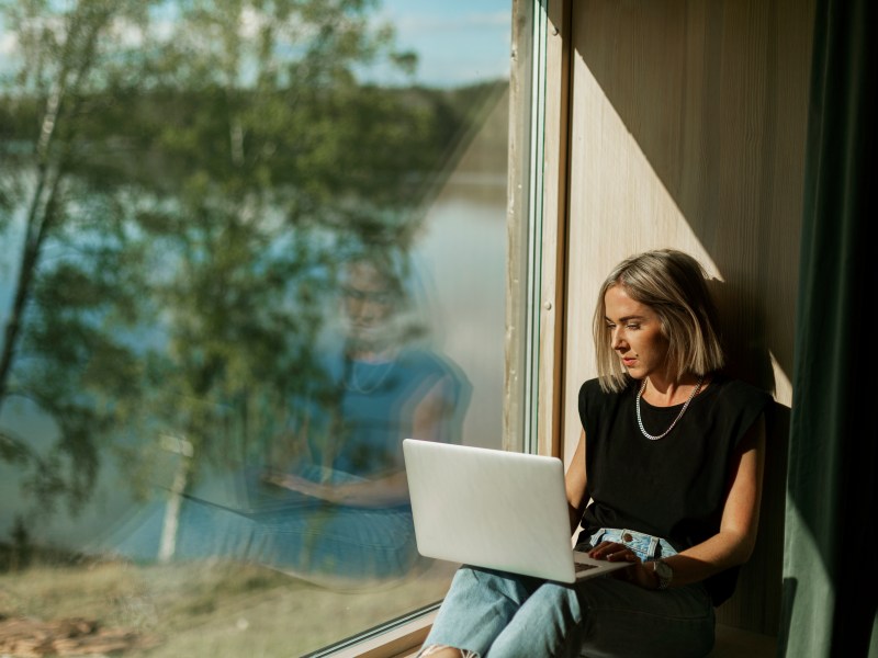 Eine Frau mit blonden Bob sitzt auf einer Fensterbank und arbeitet am Laptop.