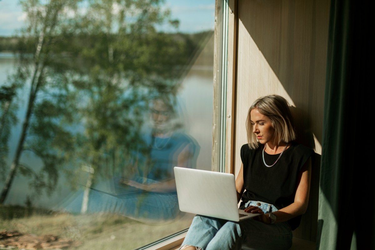 Eine Frau mit blonden Bob sitzt auf einer Fensterbank und arbeitet am Laptop.