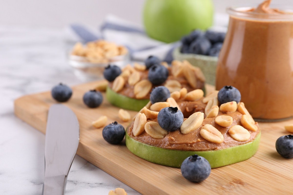 Apfel-Erdnussbutter-Bites auf einem Holzbrett. Sie sind mit Blaubeeren und ErdnÃ¼ssen garniert.