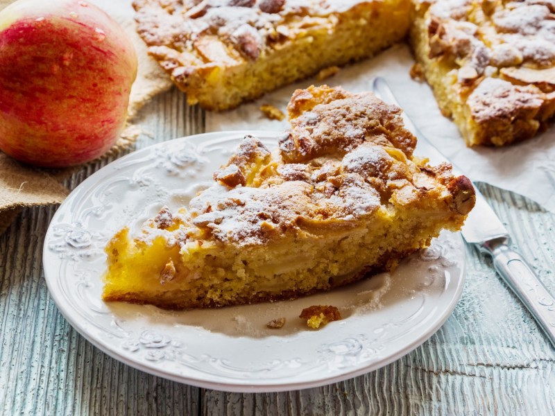 Ein StÃ¼ck Apfelkuchen ohne Mehl auf einem Teller, daneben der angeschnittene Kuchen und ein Apfel sowie Besteck.