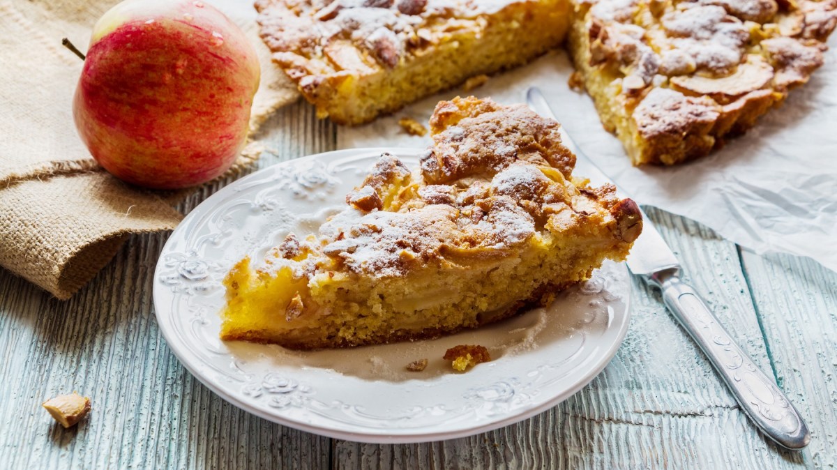 Ein StÃ¼ck Apfelkuchen ohne Mehl auf einem Teller, daneben der angeschnittene Kuchen und ein Apfel sowie Besteck.