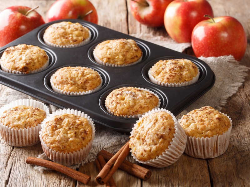 Ein Muffinblech mit Apfelmus-Muffins, drumherum weitere Muffins und frische Ã„pfel sowie Zimtstangen.