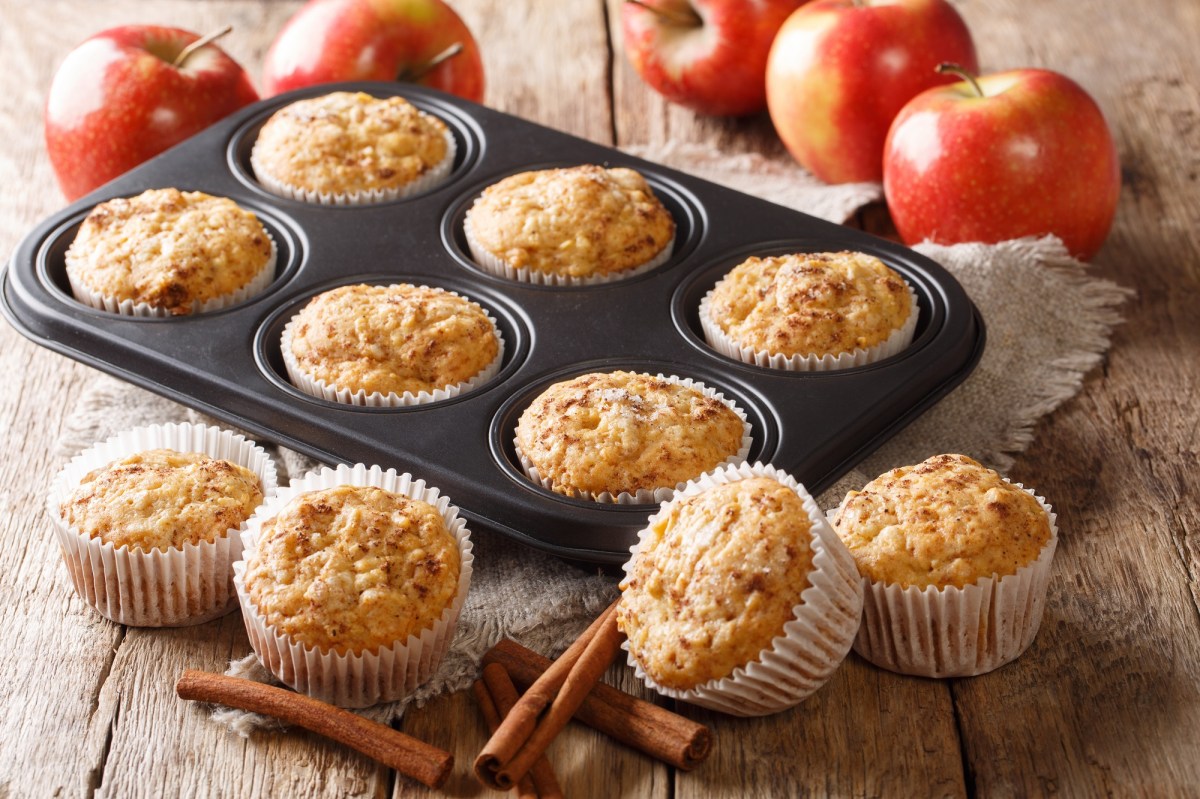 Ein Muffinblech mit Apfelmus-Muffins, drumherum weitere Muffins und frische Ã„pfel sowie Zimtstangen.