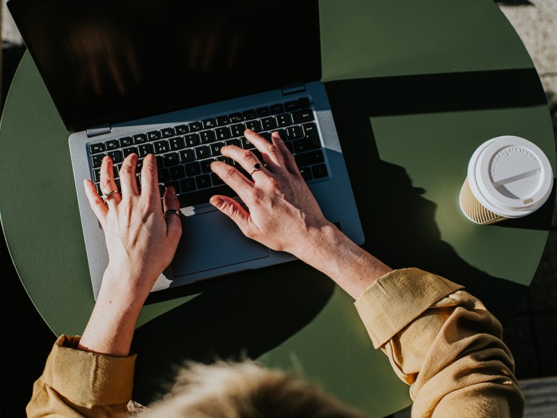 Eine Frau arbeitet drauÃŸen an ihrem Laptop. Neben dem PC steht ein Kaffee to go.