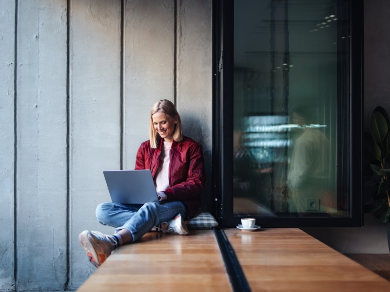 Eine junge Frau in einem lÃ¤ssigen Outfit lÃ¤chelt, wÃ¤hrend sie ihren Laptop an einem gemÃ¼tlichen Fensterplatz im CafÃ© benutzt.