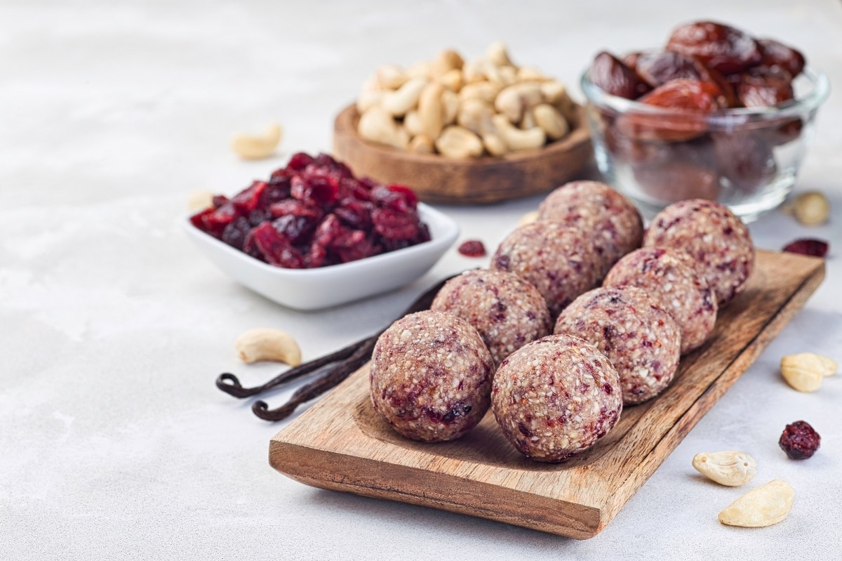 Cranberry Energy Balls auf einem Holzbrett. Dahinter die Zutaten Cranberrys, Datteln und Cashewkerne.