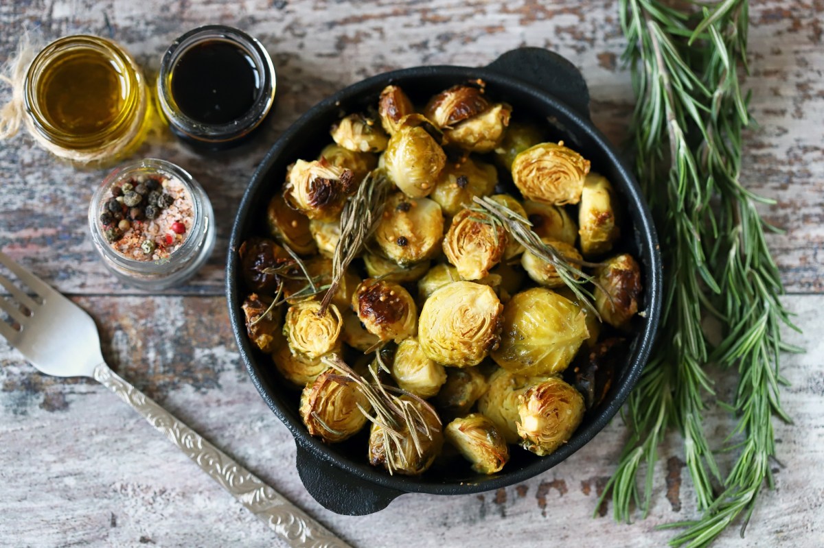 Gebackener Rosenkohl mit Balsamico und Ahornsirup in einer gusseisernen Form in der Draufsicht, daneben GlÃ¤ser mit Zutaten und Rosmarin.