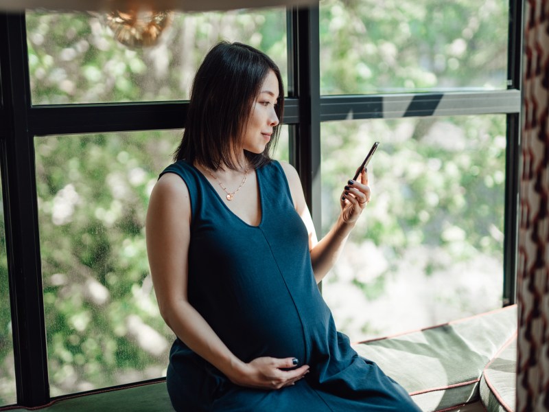 Aufnahme einer asiatischen schwangeren Frau, die ihr Mobiltelefon benutzt und eine Hand auf ihren Babybauch legt.