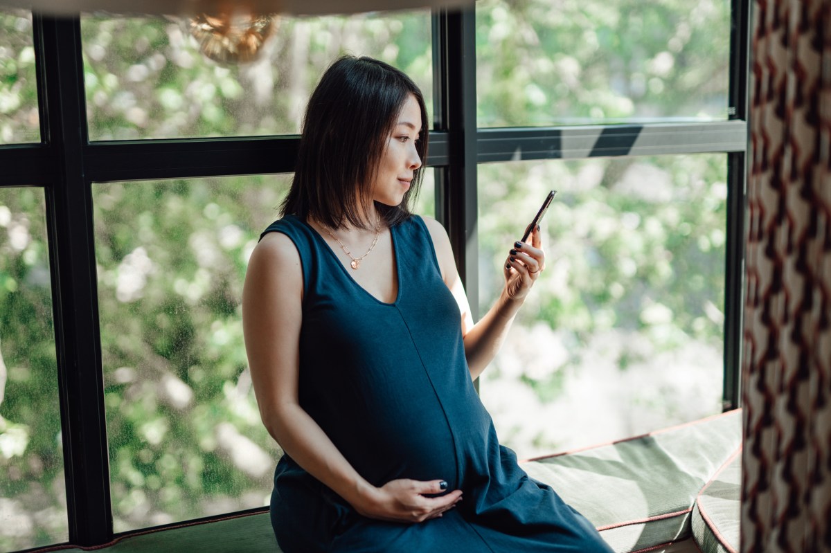 Aufnahme einer asiatischen schwangeren Frau, die ihr Mobiltelefon benutzt und eine Hand auf ihren Babybauch legt.