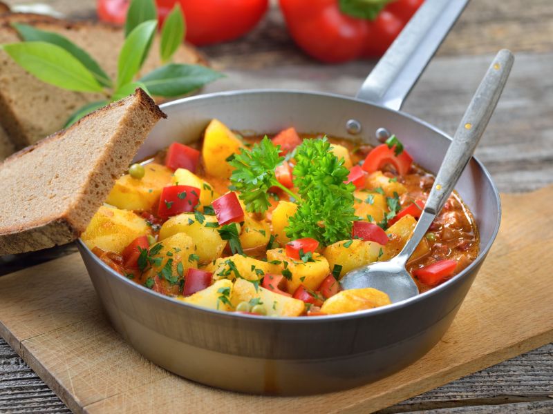 Paprika-Kartoffel-Topf, serviert in einer Pfanne. Ein LÃ¶ffel steckt im Essen, eine Scheibe Brot liegt daneben.