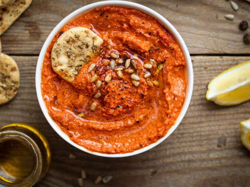 Eine Schale mit Paprika-Tomaten-Hummus. Daneben liegen frische Zutaten.