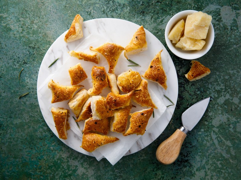 Pikante BlÃ¤tterteig-Happen auf einem weiÃŸen Teller, der auf dunkelgrÃ¼ner Unterlage steht. Daneben eine Schale mit Parmesan und ein Parmesan-Messer.