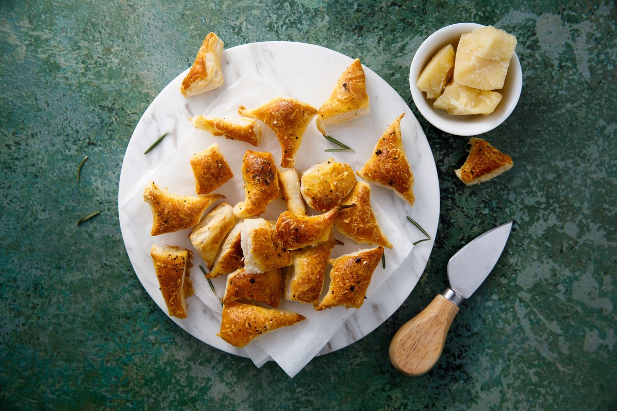 Pikante BlÃ¤tterteig-Happen auf einem weiÃŸen Teller, der auf dunkelgrÃ¼ner Unterlage steht. Daneben eine Schale mit Parmesan und ein Parmesan-Messer.