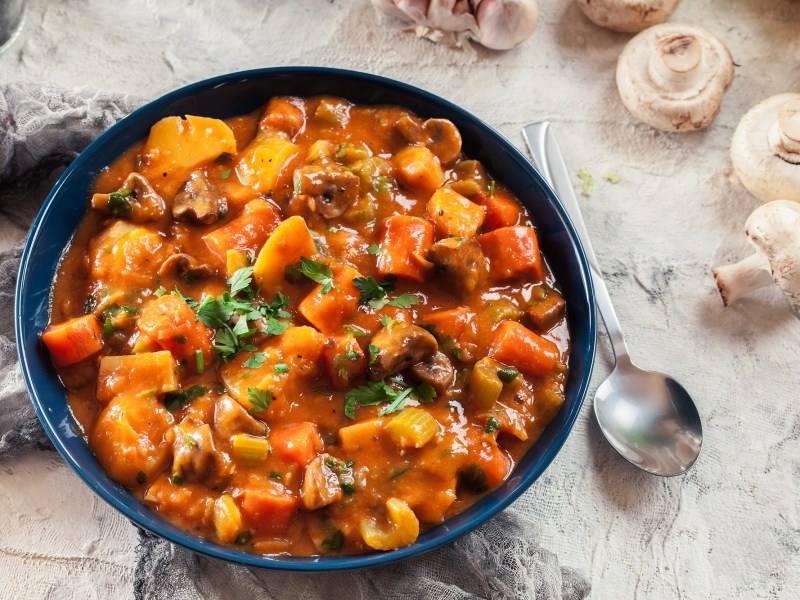 Ein Teller Pilzgulasch mit MÃ¶hren und Sellerie, daneben ein LÃ¶ffel und Pilze, Draufsicht.