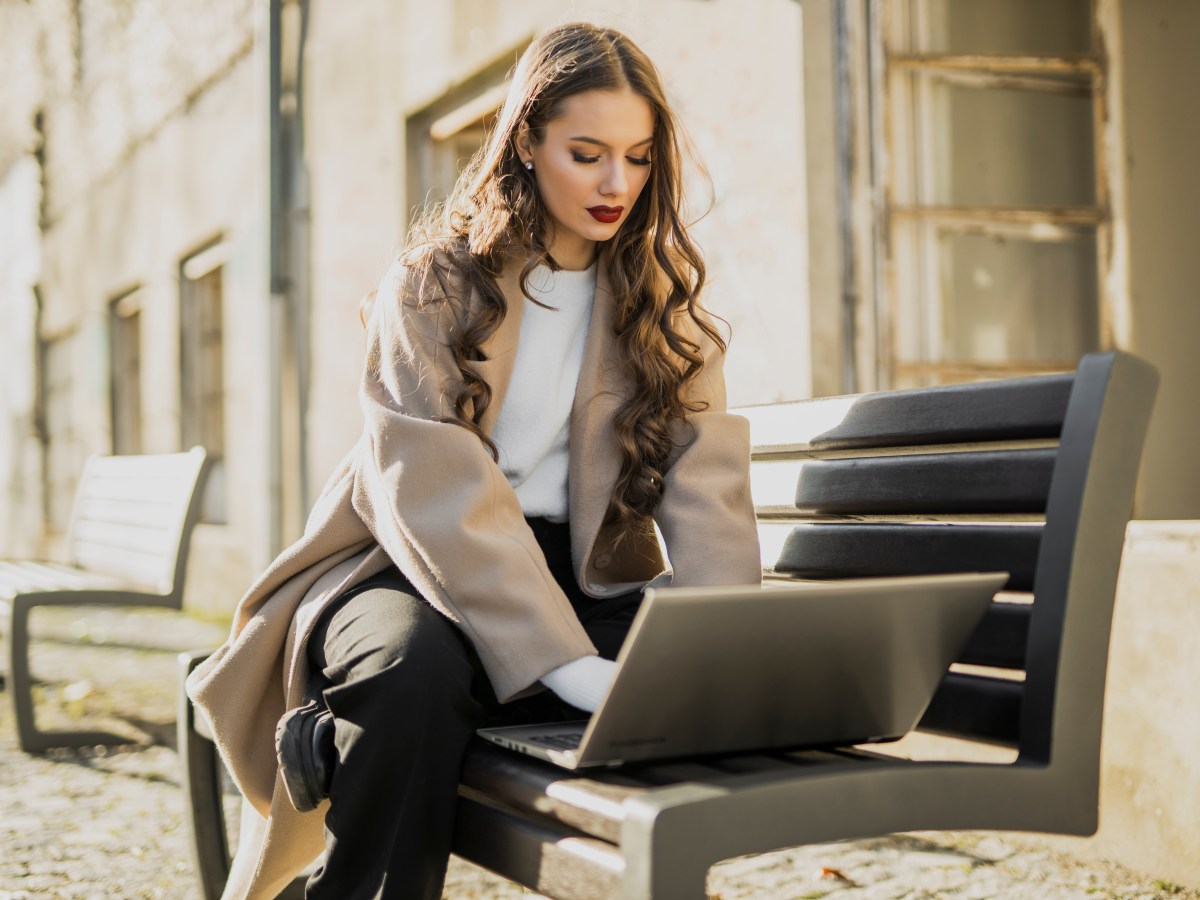 Junge Frau im Trenchcoat sitzt auf einer Bank und arbeitet am Laptop.