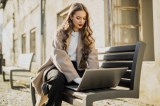 Junge Frau im Trenchcoat sitzt auf einer Bank und arbeitet am Laptop.