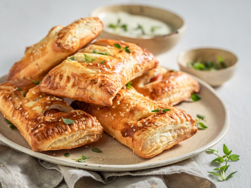 mehrere Spinat-BlÃ¤tterteig-Taschen mit Feta auf einem Teller, im Hintergrund eine Schale mit Dip und eine mit KrÃ¤utern.