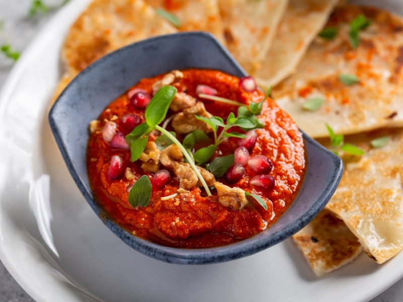 Eine Schale Tomate-Walnuss-Aufstrich, garniert mit WalnÃ¼ssen, KrÃ¤utern und Granatapfelkernen, auf einem Teller, auf dem auch dreieckige Fladenbrote liegen, Nahaufnahme.