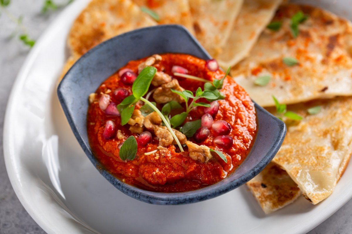 Eine Schale Tomate-Walnuss-Aufstrich, garniert mit WalnÃ¼ssen, KrÃ¤utern und Granatapfelkernen, auf einem Teller, auf dem auch dreieckige Fladenbrote liegen, Nahaufnahme.