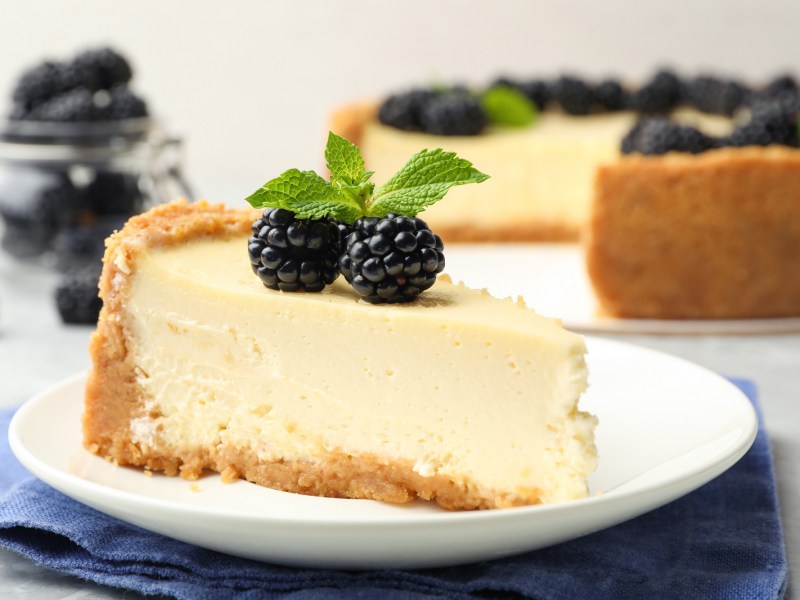 Ein StÃ¼ck veganer KÃ¤sekuchen auf einem Teller, garniert mit Minze und Brombeeren, dahinter unscharf der angeschnittene Kuchen.