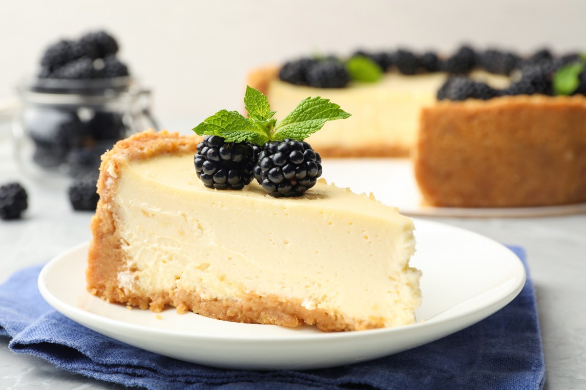 Ein StÃ¼ck veganer KÃ¤sekuchen auf einem Teller, garniert mit Minze und Brombeeren, dahinter unscharf der angeschnittene Kuchen.