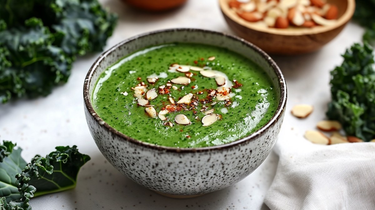 Eine SchÃ¼ssel mit GrÃ¼nkohl-Kokos-Suppe garniert mit gehackten Mandeln und Chilipulver. Die SchÃ¼ssel steht auf einer hellen ArbeitsflÃ¤che. Ringsum liegen GrÃ¼nkohlblÃ¤tter. AuÃŸerdem ist im Hintergrund eine Schale mit Mandeln angedeutet. Es liegen zusÃ¤tzlich ein paar einzelne Mandeln verstreut.