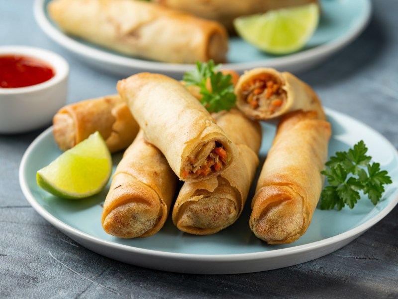 Kimchi-FrÃ¼hlingsrollen liegen auf einem hellblauen Teller. Sie sind mit einem LimettenstÃ¼ck und frischer Petersilie garniert. Im Hintergrund steht eine Schale mit SoÃŸe, sowie ein weiterer Teller FrÃ¼hlingsrollen.